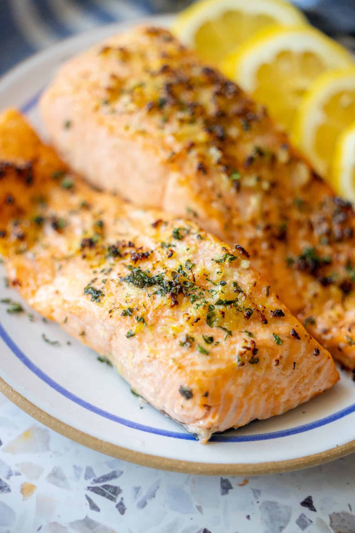 A pair of succulent baked salmon fillets, generously seasoned with herbs and spices, presented on a plate. Garnished with three bright lemon slices in the background, this dish is a testament to a quick and easy air fryer salmon preparation, ideal for a keto-friendly meal.