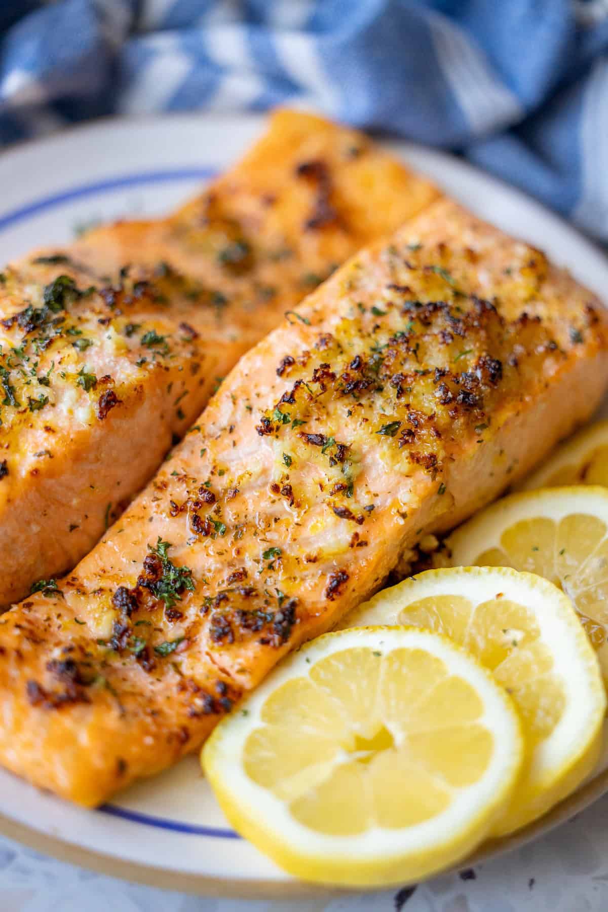 A striking overhead shot of two perfectly cooked air fryer salmon fillets, adorned with a sprinkle of fresh herbs, nestled beside vibrant lemon slices on a crisp white plate. A textured blue and white cloth subtly complements the dish, emphasizing the effortless elegance of this Easy Air Fryer Lemon Garlic Salmon Recipe.