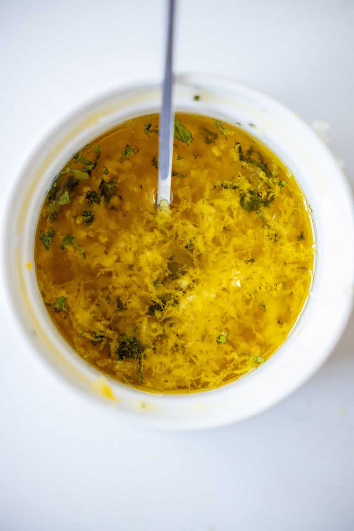 A close-up top view of a bowl containing a yellow mixture of oil, herbs, and grated zest with a spoon inside—perfect for making an Easy Air Fryer Lemon Garlic Salmon Recipe.