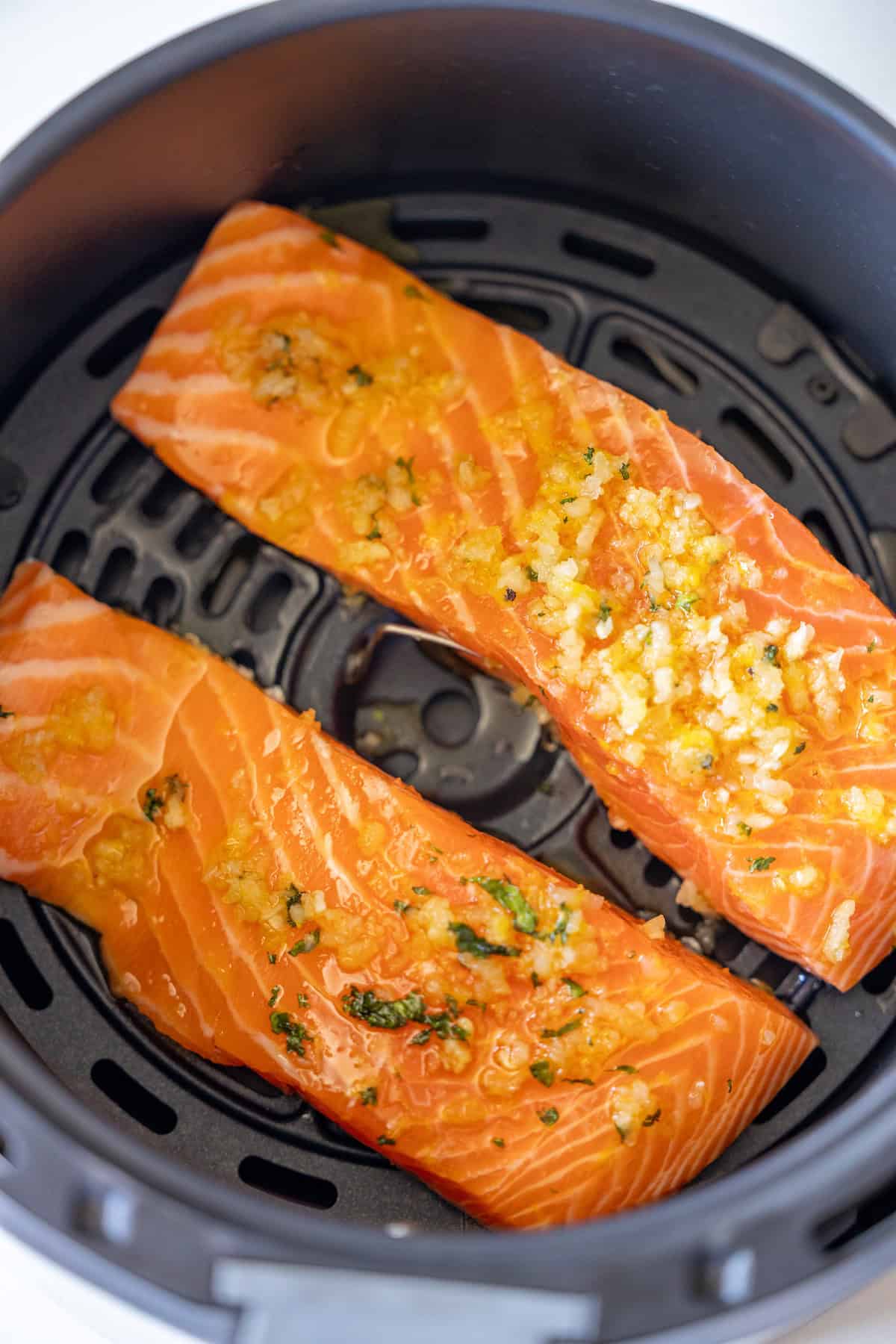 Two raw salmon fillets topped with chopped garlic and herbs placed inside an air fryer basket, ready to be cooked—an Easy Air Fryer Lemon Garlic Salmon Recipe perfect for learning how to air fry salmon effortlessly.