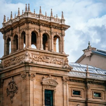 A stone building with intricate architectural details and a tower featuring multiple arches and ornate carvings under a partially cloudy sky, akin to the historic charm you'd find in a travel guide to San Sebastian.