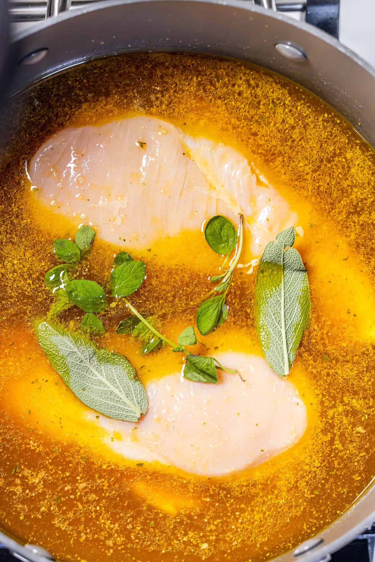 Two raw chicken breasts in a pot with orange-colored broth, garnished with fresh sage and oregano—perfect for an easy boiled chicken recipe or meal prep boiled chicken recipe.