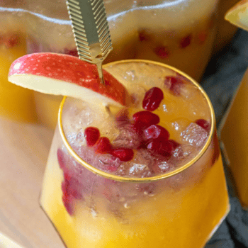 A yellow drink served in a glass with ice, pomegranate seeds, and a red apple slice garnish on the rim, featuring a gold stirrer. This delightful concoction is reminiscent of a pumpkin puree cocktail.