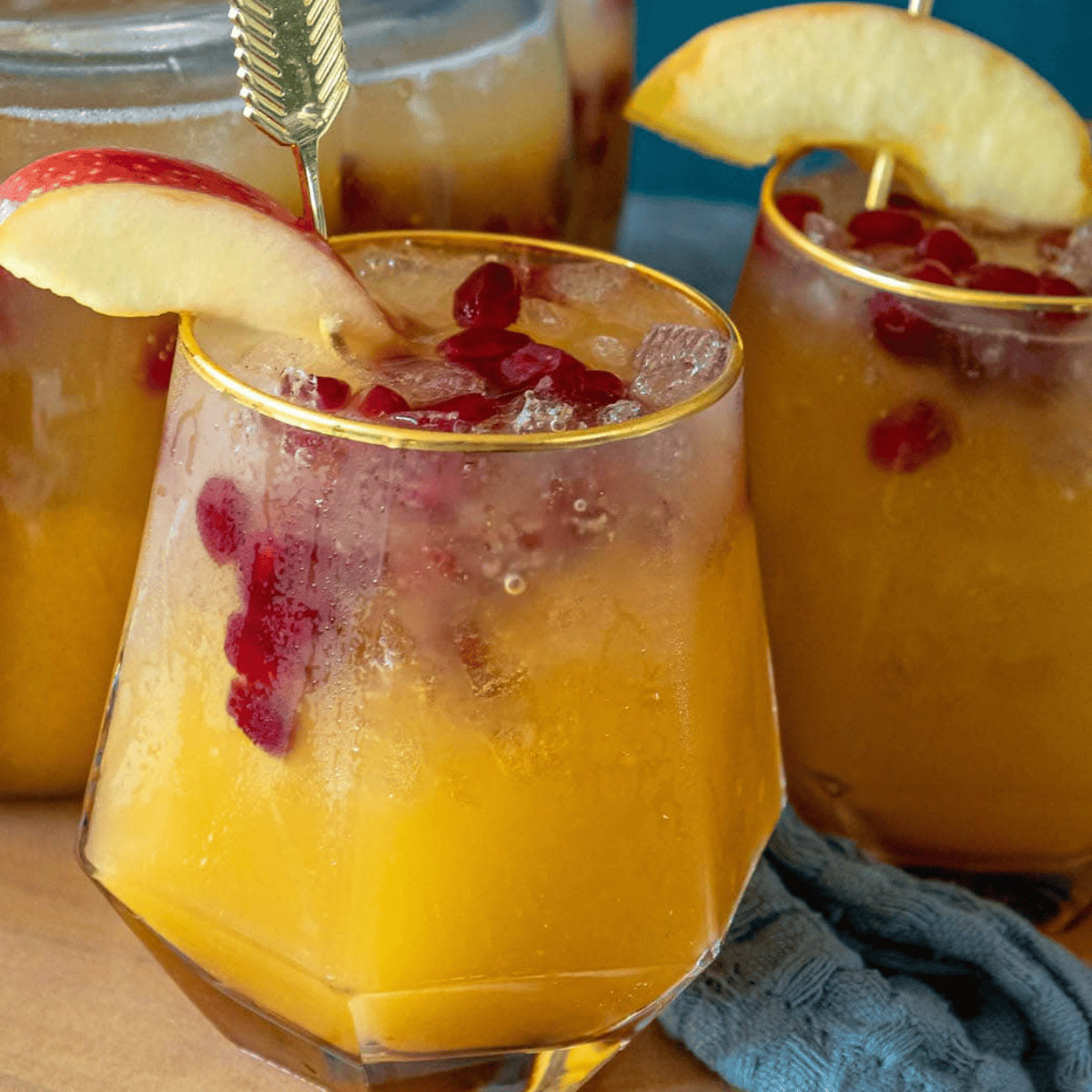 Two glasses filled with a yellow-orange pumpkin punch cocktail, garnished with red pomegranate seeds and apple slices, with a pitcher of the same festive drink in the background.