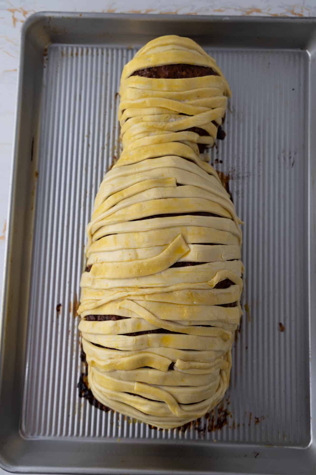 A pastry-wrapped loaf resembling a mummy shape, placed on a baking sheet. This delectable mummy meatloaf is covered with layered dough strips.