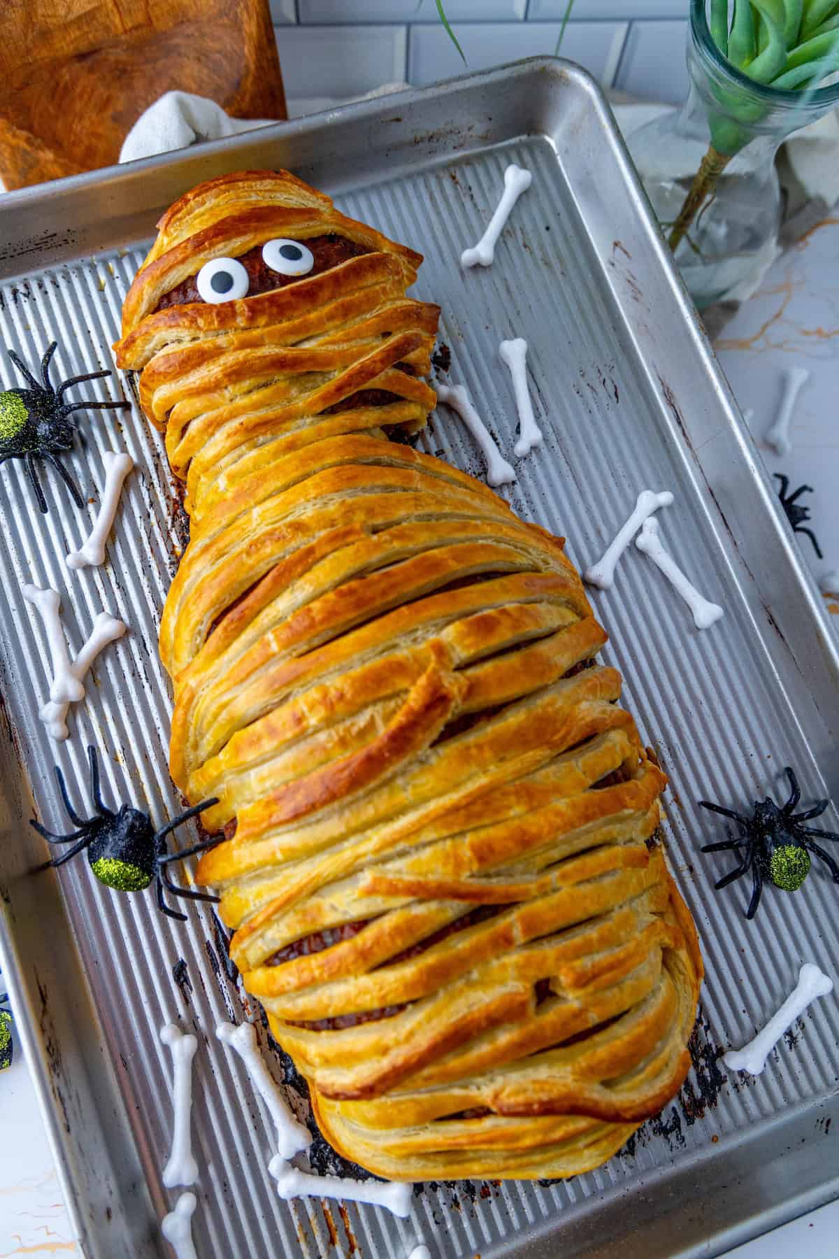 A tray features a mummy-shaped meatloaf with googly eyes, surrounded by decorative bones and spiders.