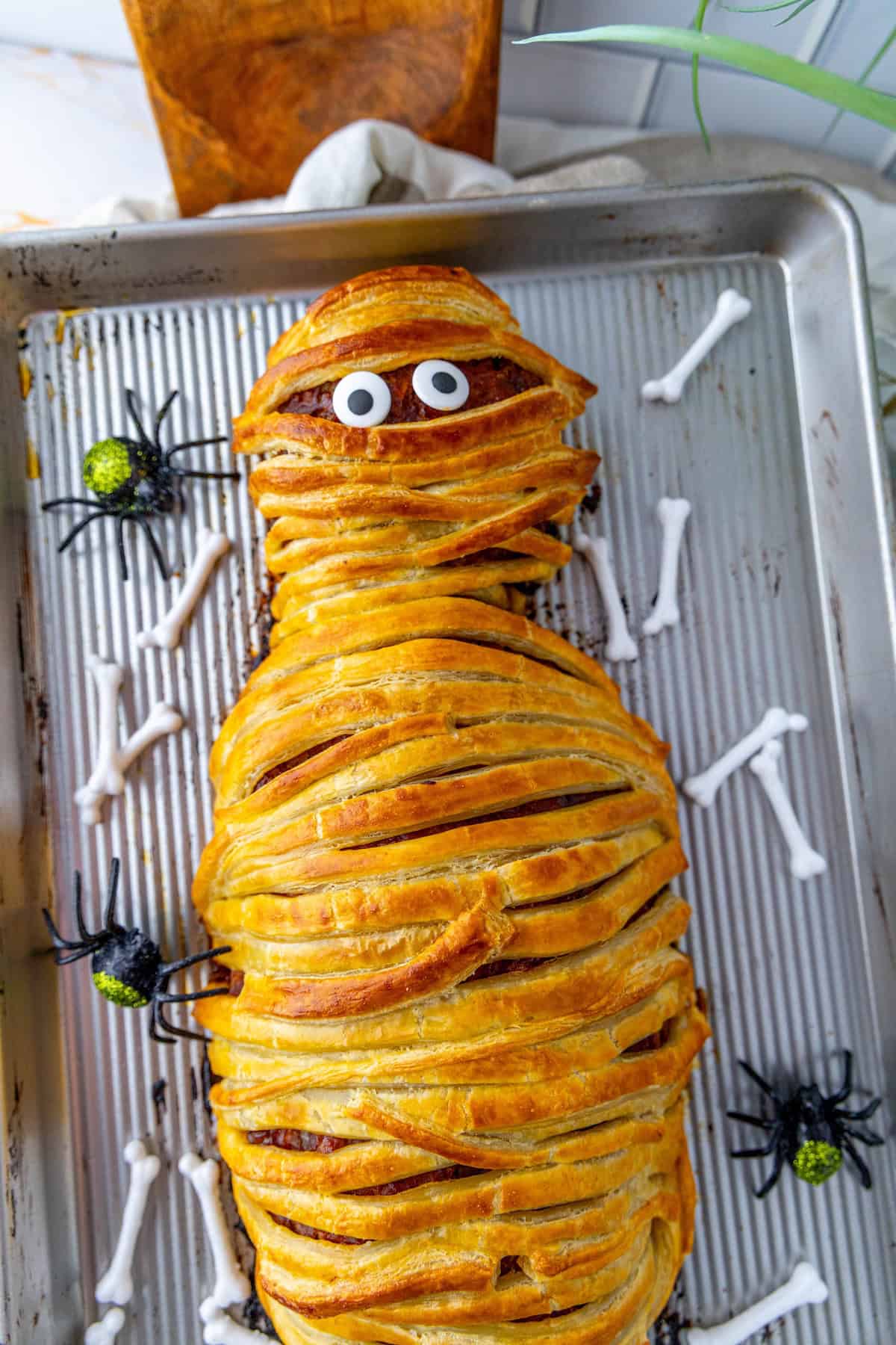 A mummy meatloaf wrapped in pastry, decorated with candy eyes. Plastic spiders and bone-shaped props are scattered around on a baking tray.