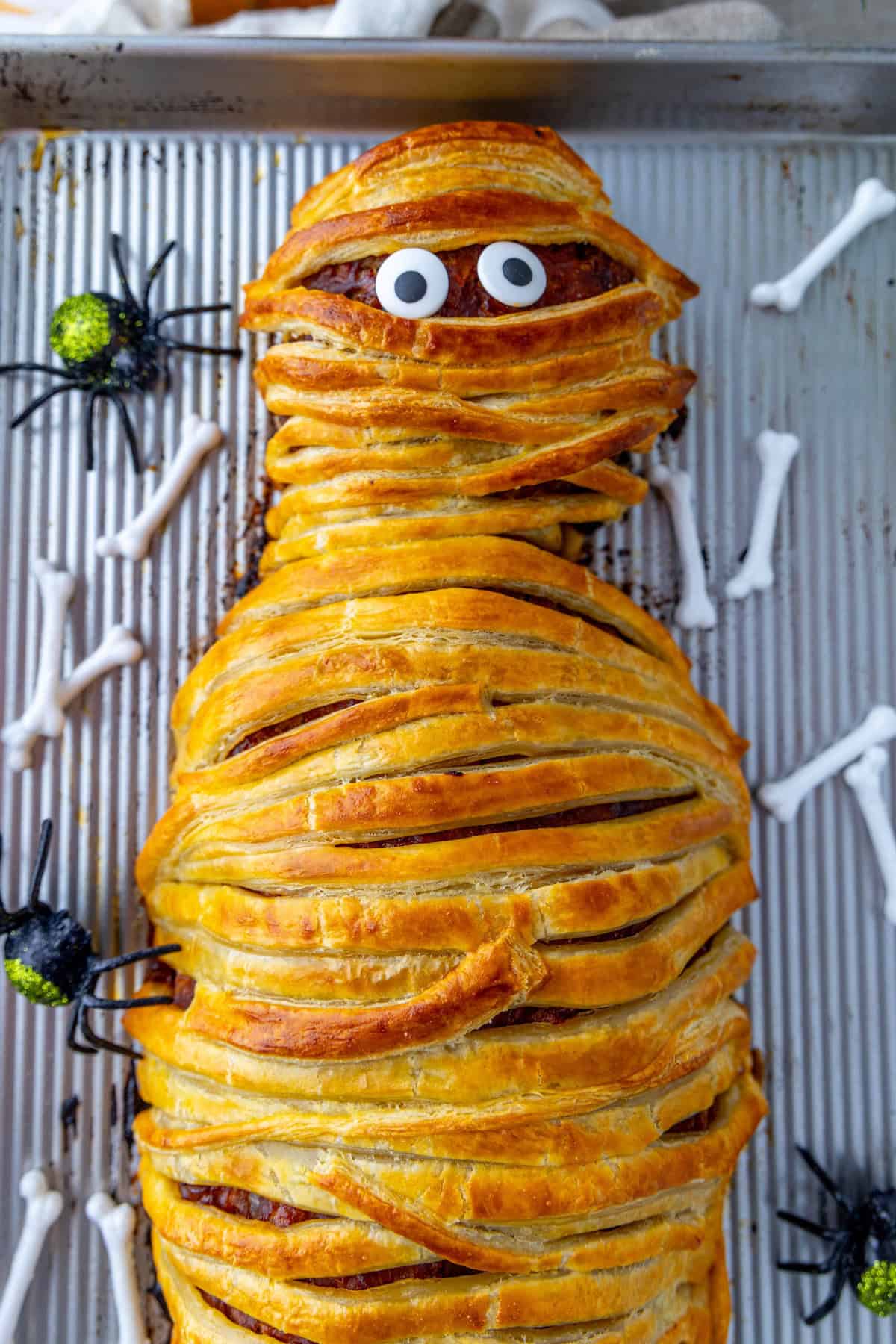 A baked dish resembling a mummy meatloaf, made from wrapped pastry with googly eyes, is placed on a baking sheet along with plastic spiders and bone decorations.