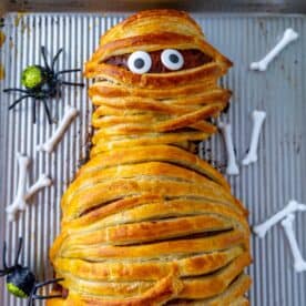 A mummy meatloaf dish wrapped in pastry with visible eyes, accompanied by plastic spiders and bones on a baking tray.