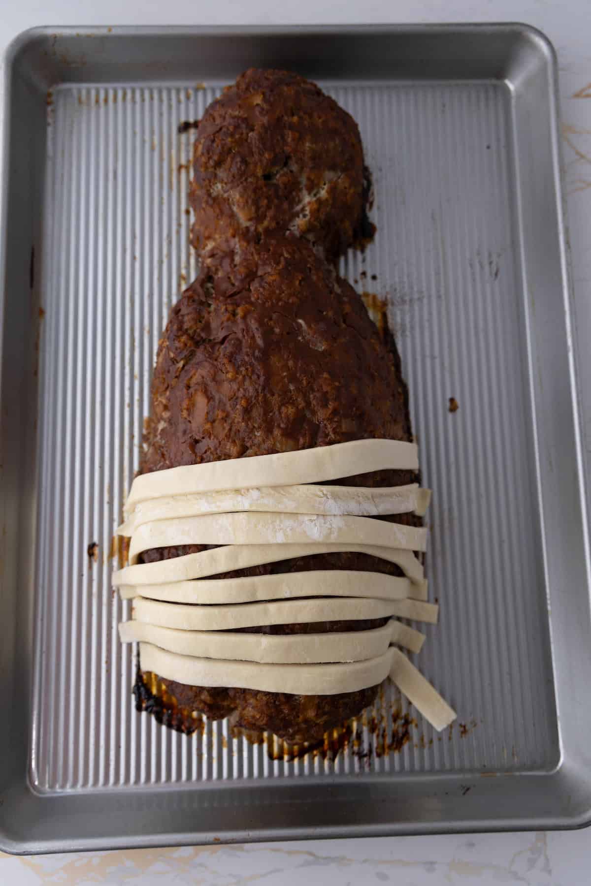 A mummy meatloaf shaped like a baby on a baking sheet, partially wrapped in strips of dough.