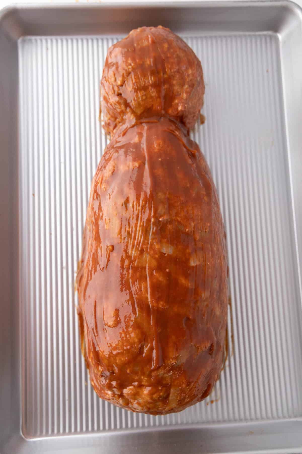 Uncooked mummy meatloaf shaped into a loaf form and coated with sauce, placed on a baking sheet with ridges.