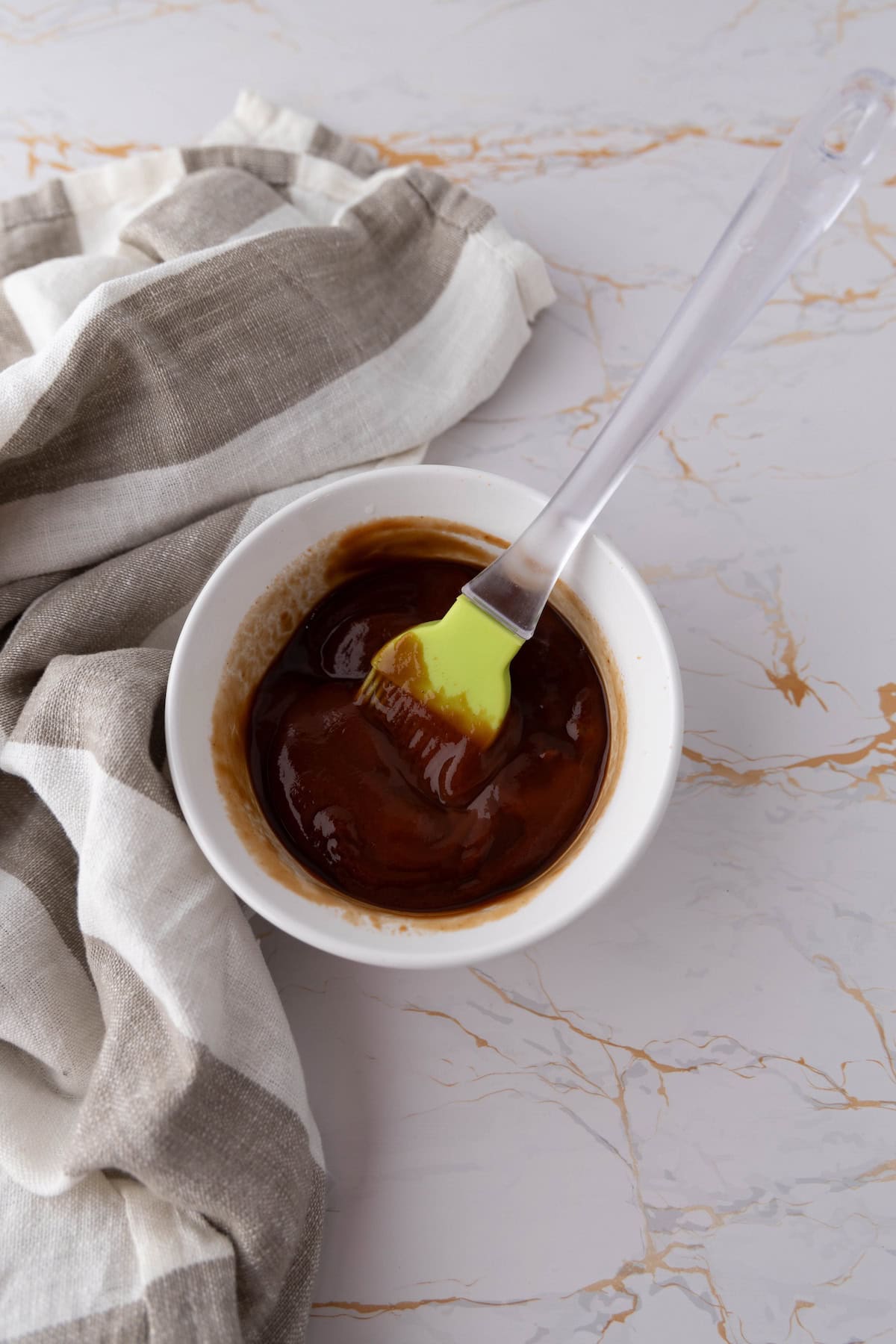 A bowl of barbecue sauce with a silicone brush in it sits on a marble surface, ready to glaze the savory mummy meatloaf next to a folded striped cloth.