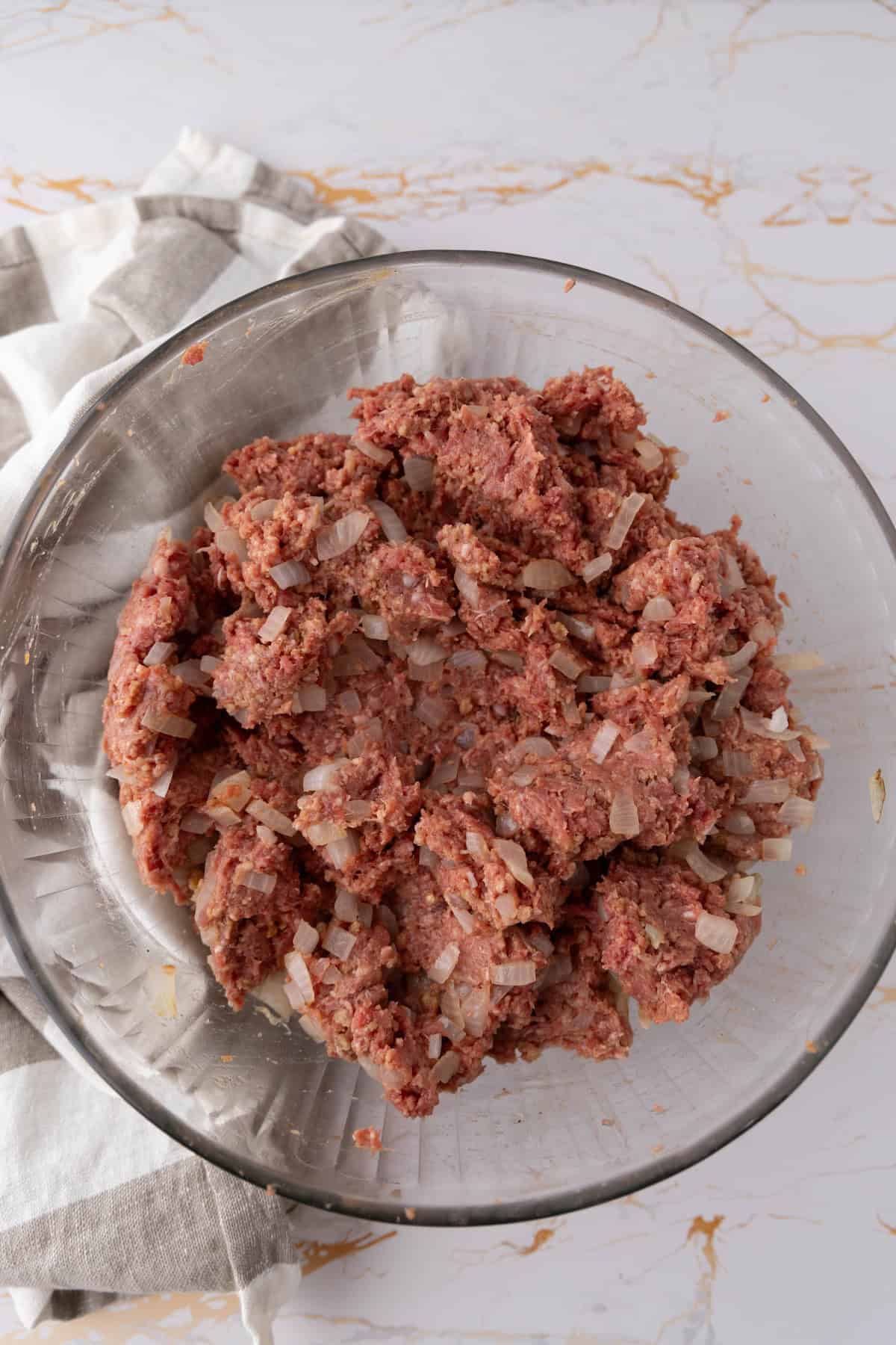 A glass bowl filled with a mixture of ground meat and chopped onions, perfect for making mummy meatloaf, sits on a folded white and gray striped cloth on a marble countertop.