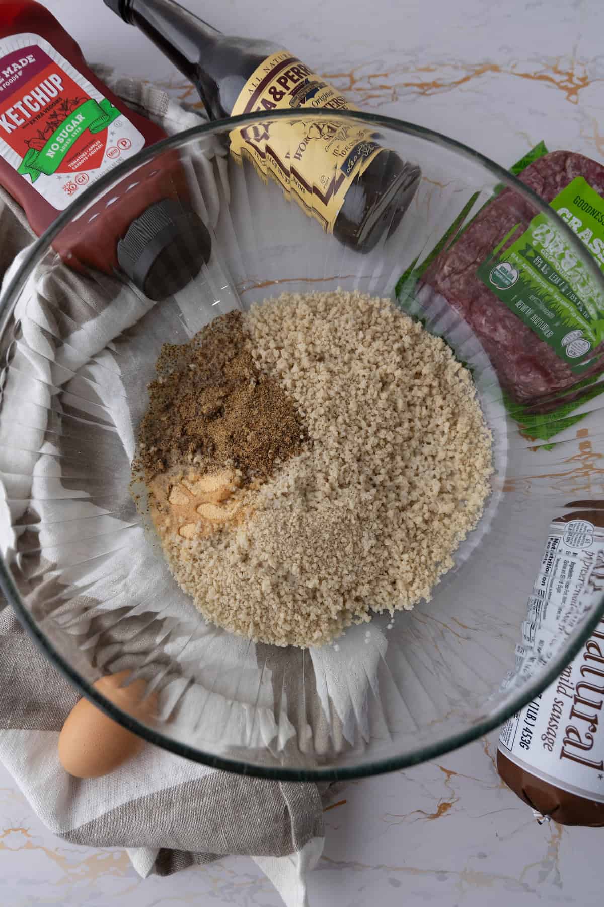 A bowl with breadcrumbs and seasoning sits amidst ketchup, Worcestershire sauce, packaged ground beef, an egg, and a package of Italian sausage—perfect for crafting a delicious mummy meatloaf.