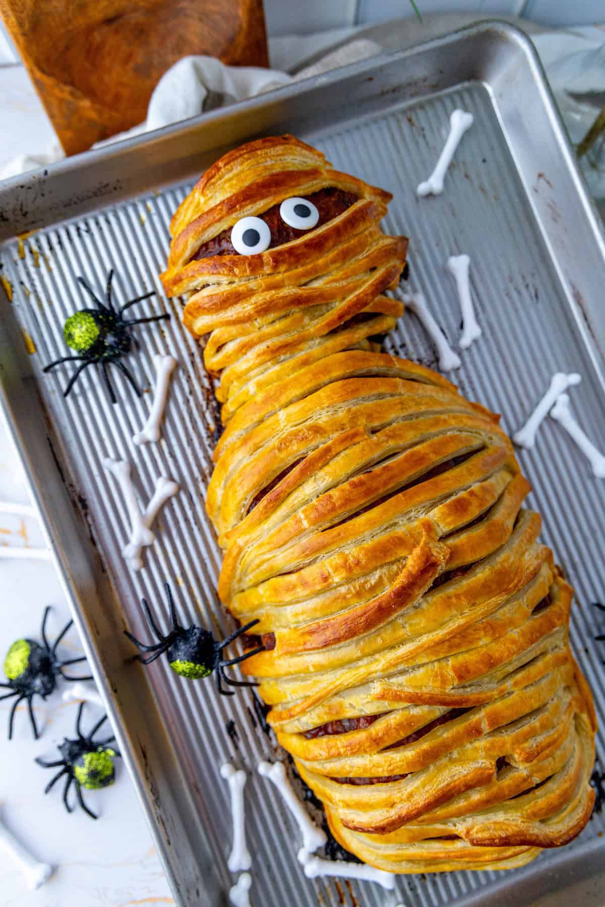 A mummy meatloaf shaped like a playful mummy with googly eyes, placed on a baking tray alongside decorative spiders and bone-shaped treats.