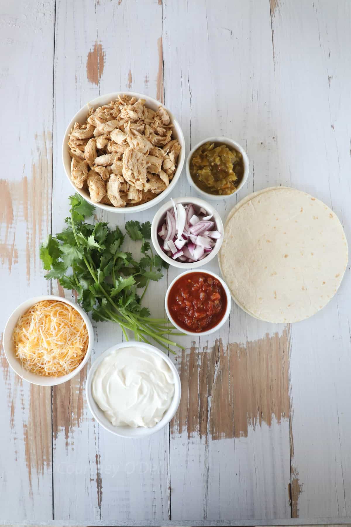 Ingredients for a meal: shredded chicken, chopped cilantro, shredded cheese, sour cream, diced onions, salsa, green chilies, and flour tortillas arranged on a wooden surface.
