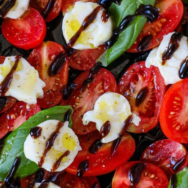 A close-up of a caprese salad with sliced tomatoes, mozzarella, basil leaves, and balsamic glaze drizzled on top.