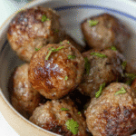 A bowl filled with several browned air fryer meatballs, garnished with small pieces of chopped fresh herbs.