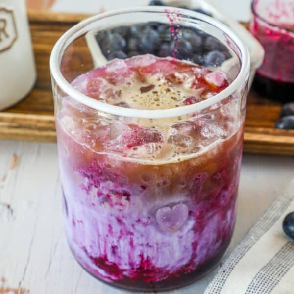 A glass of a layered drink with ice, combining purple and brown hues, appears creamy on top. Blueberries and a pink-tinted jar are in the background.