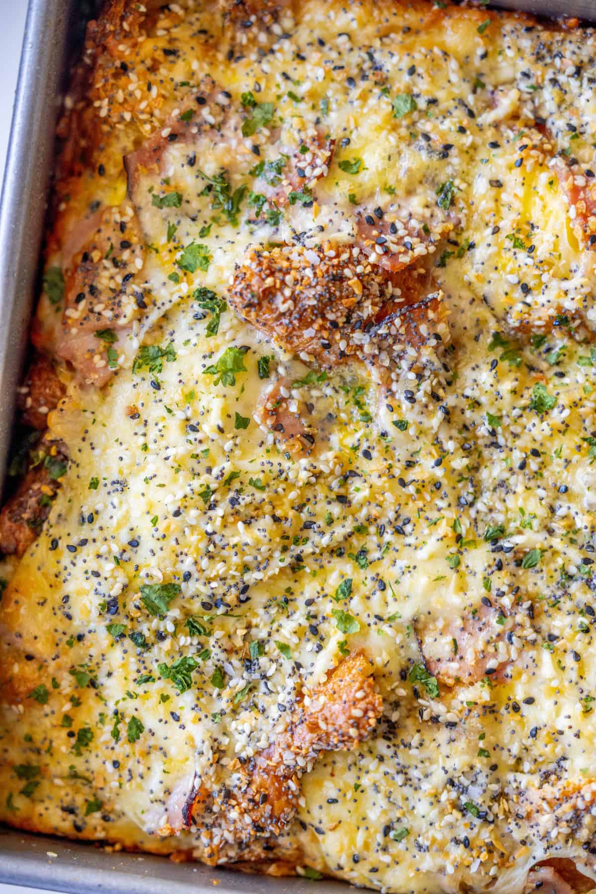 A close-up of a baked Ham and Cheese Croissant Strata topped with melted cheese, parsley, and seasoning, showing a golden crust and crispy edges. The dish appears to be a savory casserole.