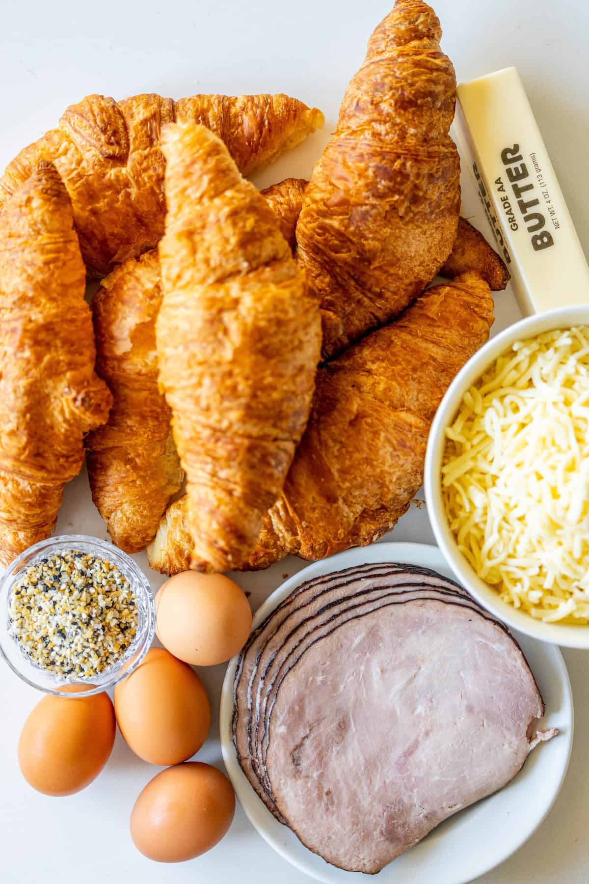 An array of ingredients, perfect for a Ham and Cheese Croissant Strata: croissants, a stick of butter, shredded cheese, sliced ham, three eggs, and a small bowl of seasoning on a white surface.