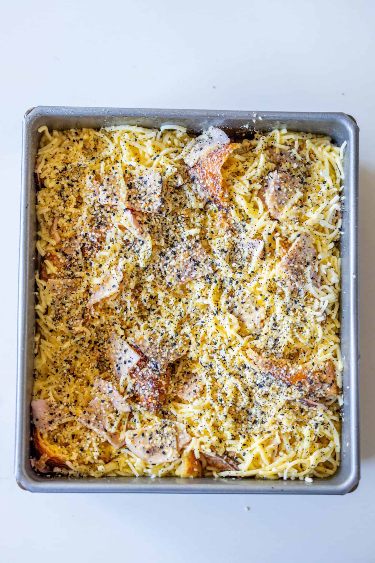 A square baking tray contains a Ham and Cheese Croissant Strata, layered with shredded cheese, pieces of cooked meat, and sprinkled with black pepper and seasonings.