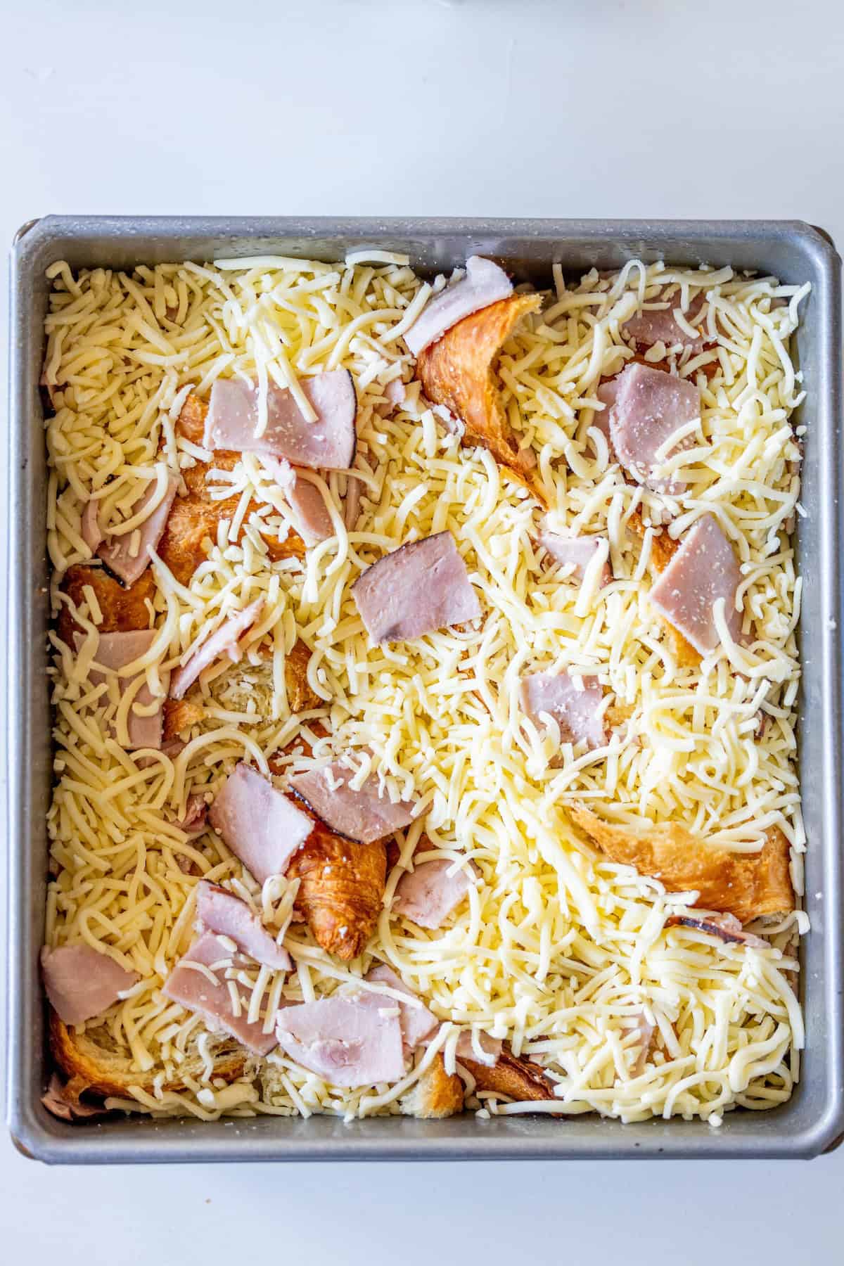 A square baking dish filled with croissant pieces, shredded cheese, and slices of ham, prepared for baking into a delicious Ham and Cheese Croissant Strata.
