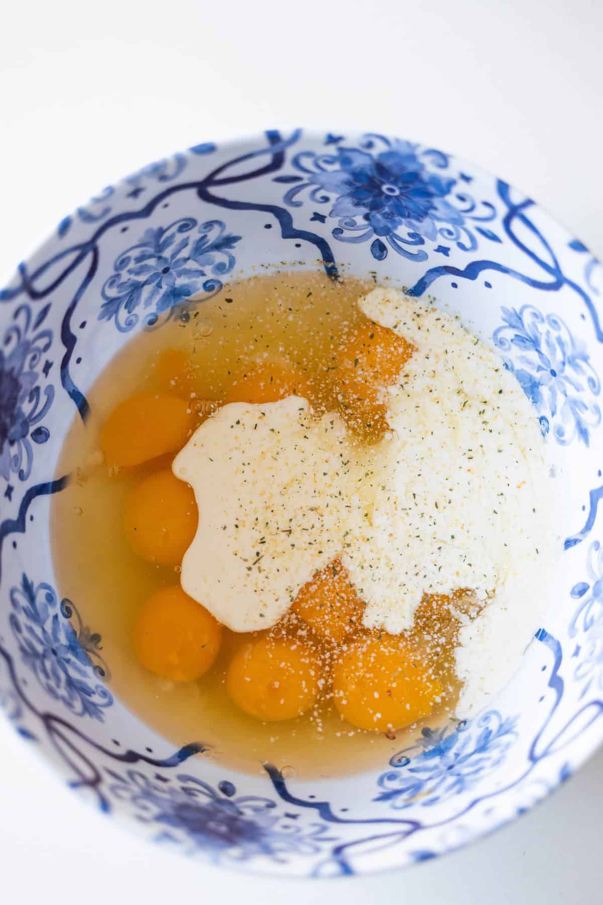 A blue and white patterned bowl contains cracked eggs, cream, specks of seasoning, ready to be transformed into a delicious Ham and Cheese Croissant Strata.