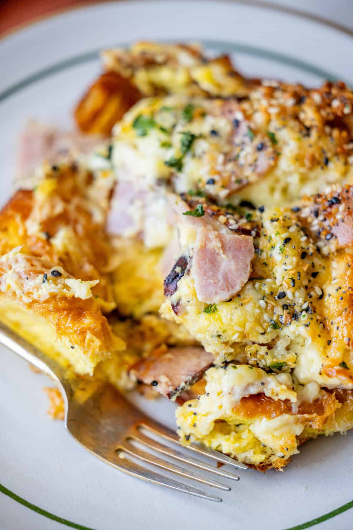 Savory Croissant Breakfast Bake 3 Close-up of a plated breakfast casserole with visible pieces of ham, scrambled eggs, and melted cheese, all nestled beside a flaky croissant. Garnished with herbs and seeds, the cheesy dish invites you in. A fork is placed near the food on the plate.