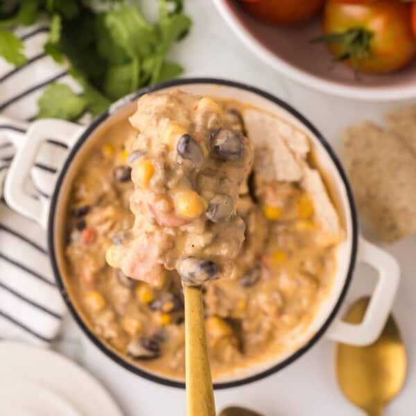 A spoonful of creamy soup with black beans, corn, and tomatoes being held above a pot resembles a Cowboy Queso recipe. A striped cloth, fresh herbs, and whole tomatoes are visible in the background.