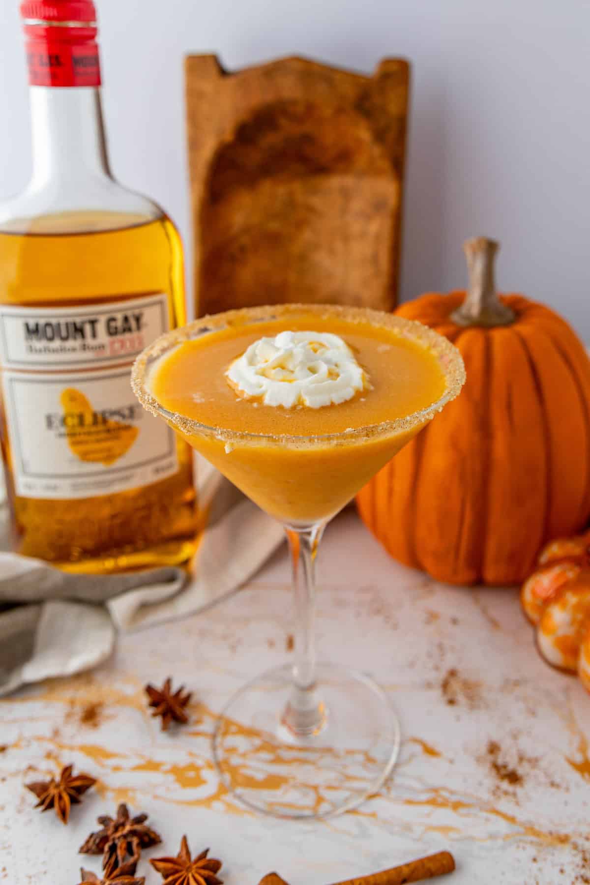 A pumpkin pie martini with whipped cream, set on a table with a bottle of Mount Gay rum, spices, and pumpkins in the background.