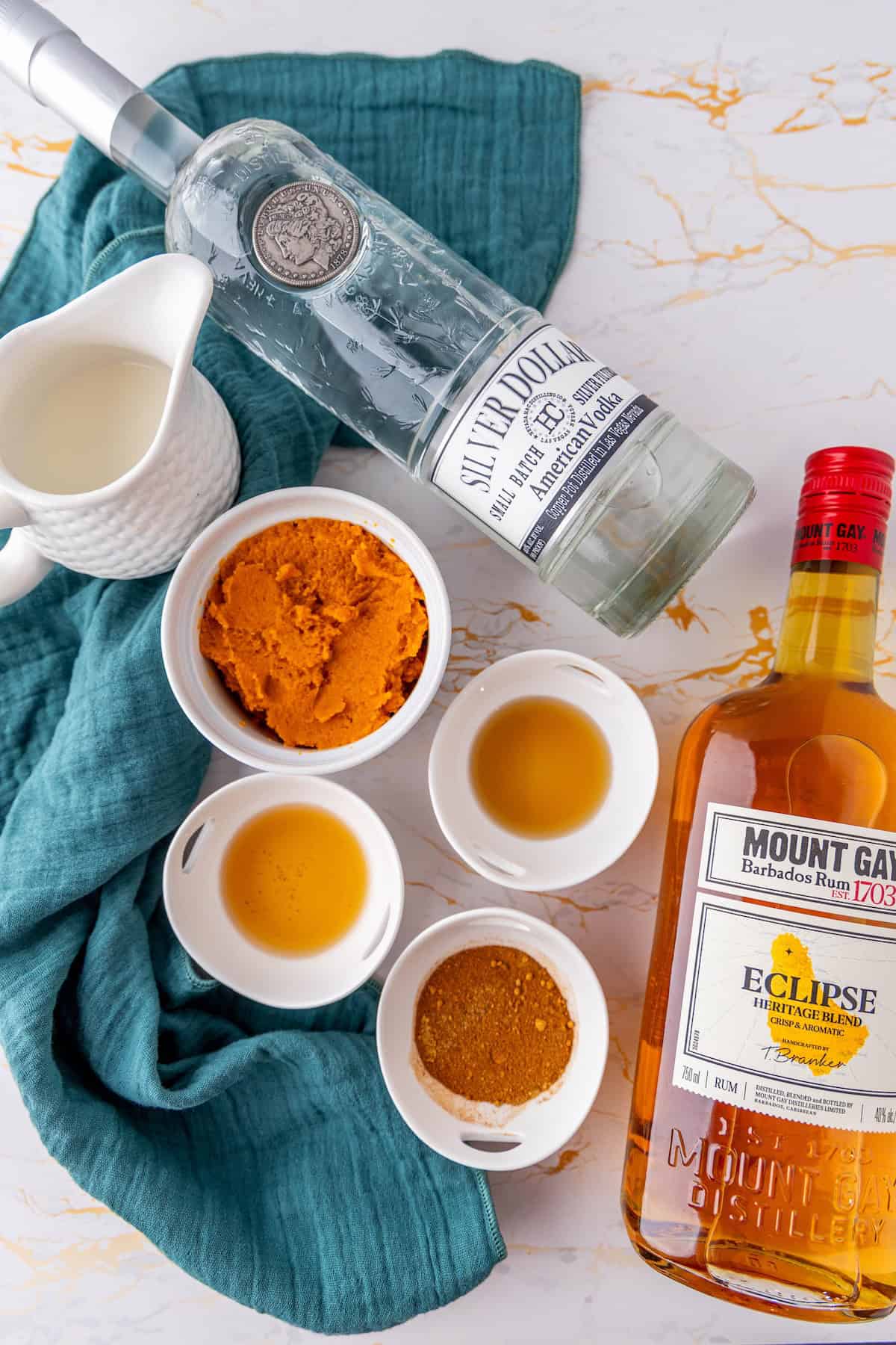 A flat lay shows ingredients including a bottle of Silver Dollar Kentucky Vodka, a bottle of Mount Gay Rum, orange puree, brown sugar, and other spices on a marble surface with a teal cloth—perfect for crafting the ultimate pumpkin pie martini.