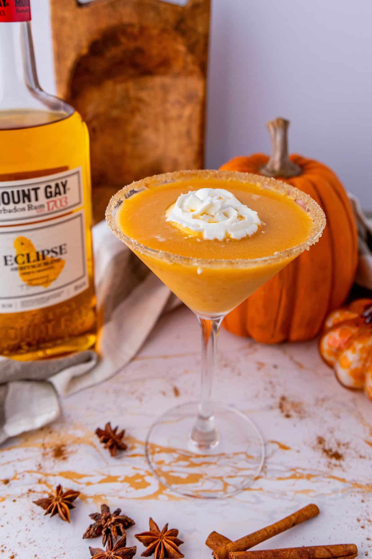 A pumpkin pie martini garnished with whipped cream in a glass with a sugar rim, surrounded by star anise, cinnamon sticks, pumpkins, and a bottle of rum on a marble surface.