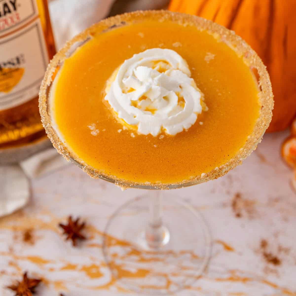 A pumpkin pie martini garnished with whipped cream is served in a sugar-rimmed glass, surrounded by autumn-themed decor.