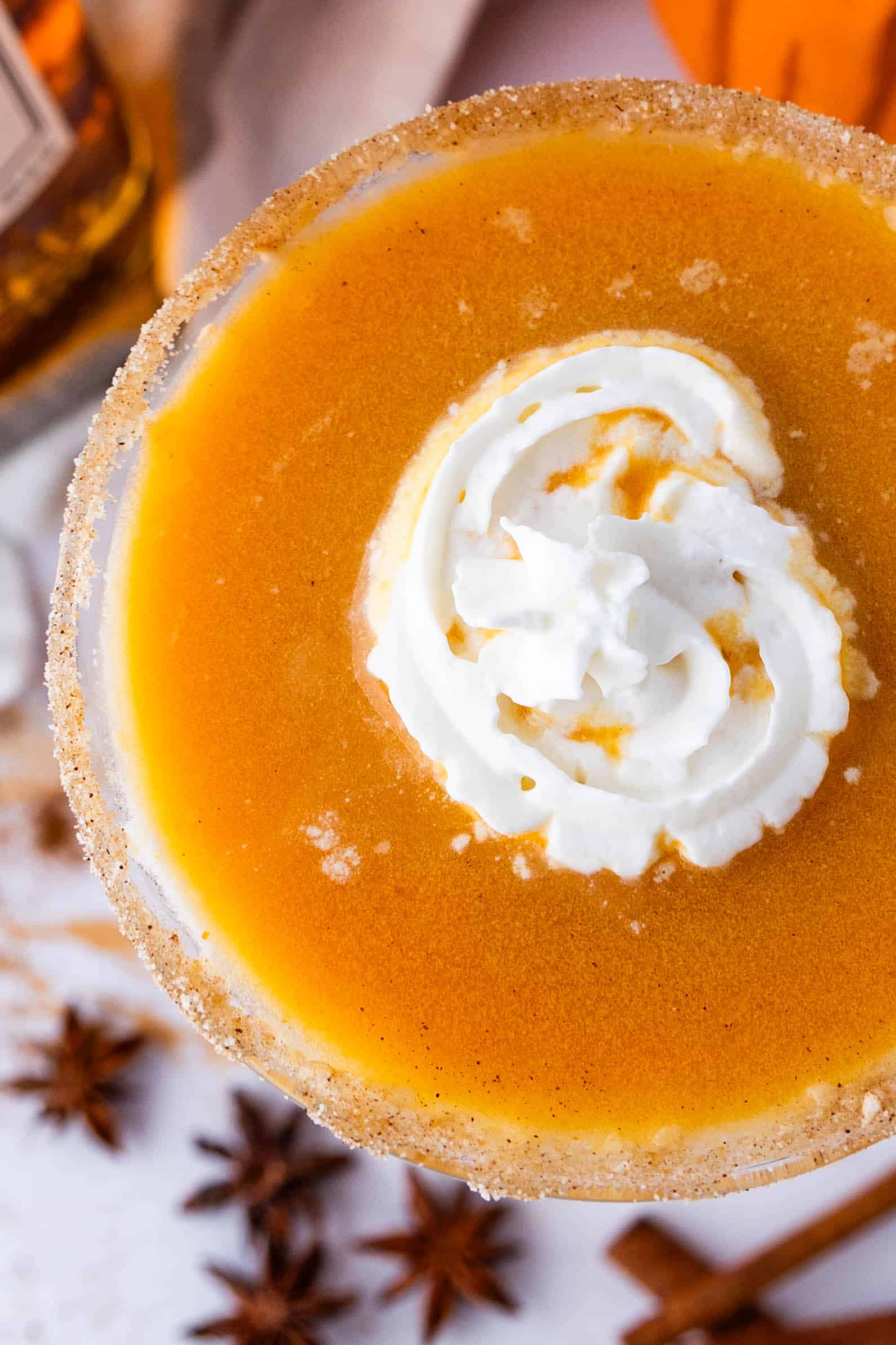 Close-up of a pumpkin pie martini topped with whipped cream, garnished with a rim coated in cinnamon sugar. Star anise and a cinnamon stick are placed nearby.