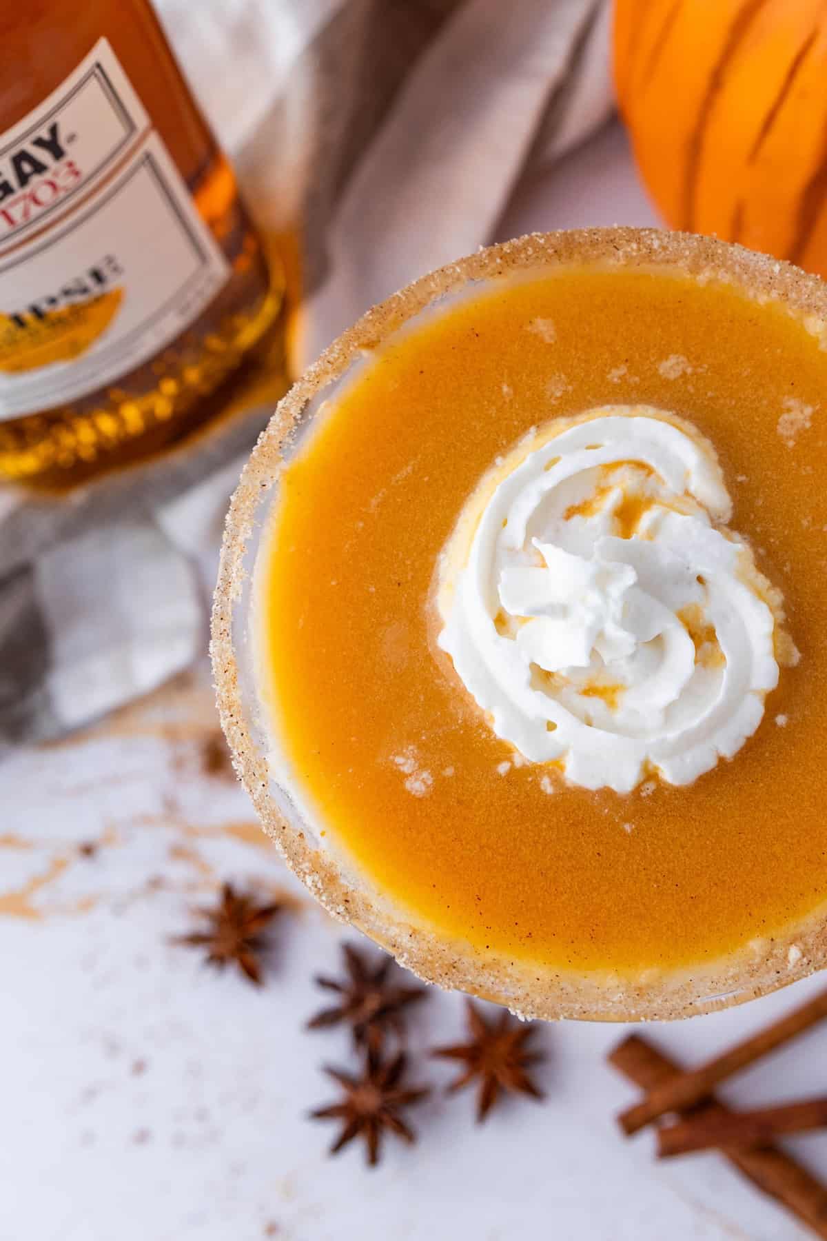 A close-up of a pumpkin pie martini topped with whipped cream. Star anise and cinnamon sticks are scattered on the table. A bottle of whiskey is partially visible in the background.