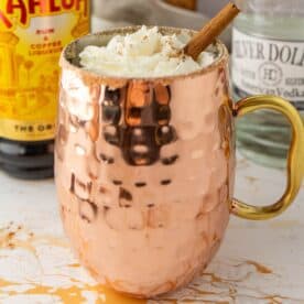 A copper mug with whipped cream and a cinnamon stick on top, surrounded by bottles of Kahlua and Silver Dollar alcohol on a white countertop.