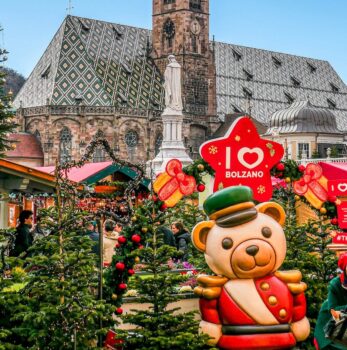 The festive Christmas market in Bolzano features charming decorated stalls, a grand teddy bear statue, and the historic cathedral beautifully framing the scene.