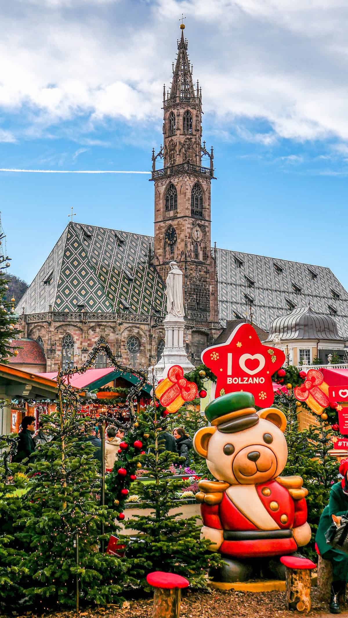 A festive Christmas market featuring decorated trees and a large bear statue stands proudly in front of Bolzano's historic church.