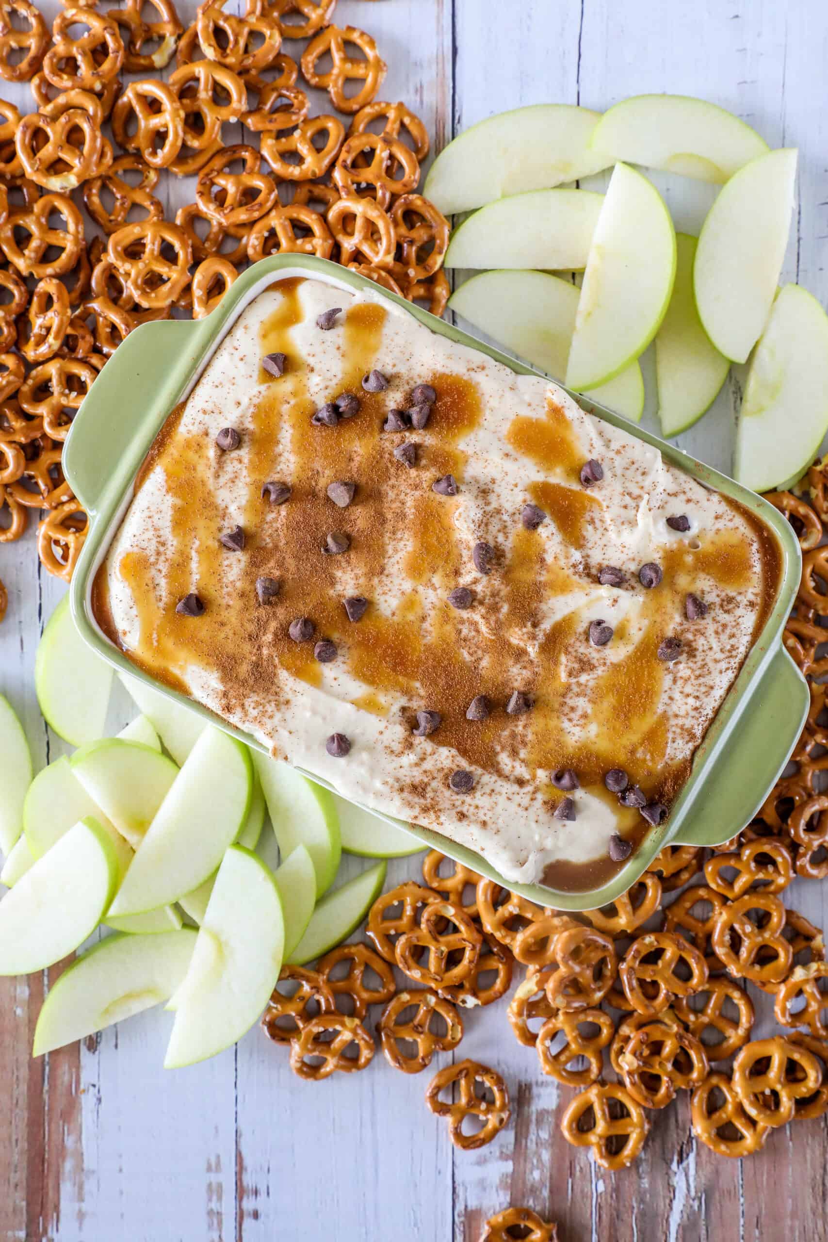 Caramel apple dip topped with chocolate chips in a dish, surrounded by sliced apples and pretzels on a wooden surface.