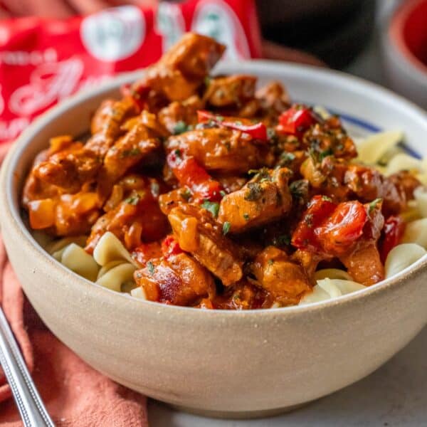 A bowl of pasta topped with a paprikash-inspired sauce featuring chicken, herbs, and red bell peppers sits enticingly on the table, with a red bag adding a splash of color in the background.