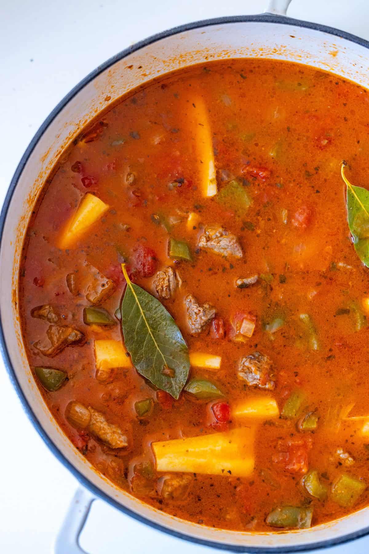 A pot contains yellow baby potatoes, minced garlic, tomato paste, diced carrots, and meat, all seasoned with green herbs for a classic beef stew.
