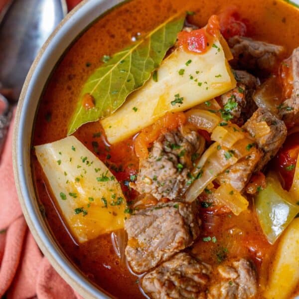 Close-up of a steaming bowl of homemade beef stew, featuring tender chunks of beef, vegetables, fresh herbs, and a fragrant bay leaf nestled in a savory, rich red broth.