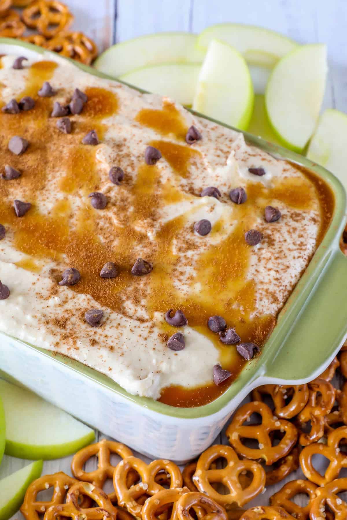 A casserole dish with creamy caramel apple dip topped with chocolate chips and cinnamon, surrounded by sliced green apples and pretzels.