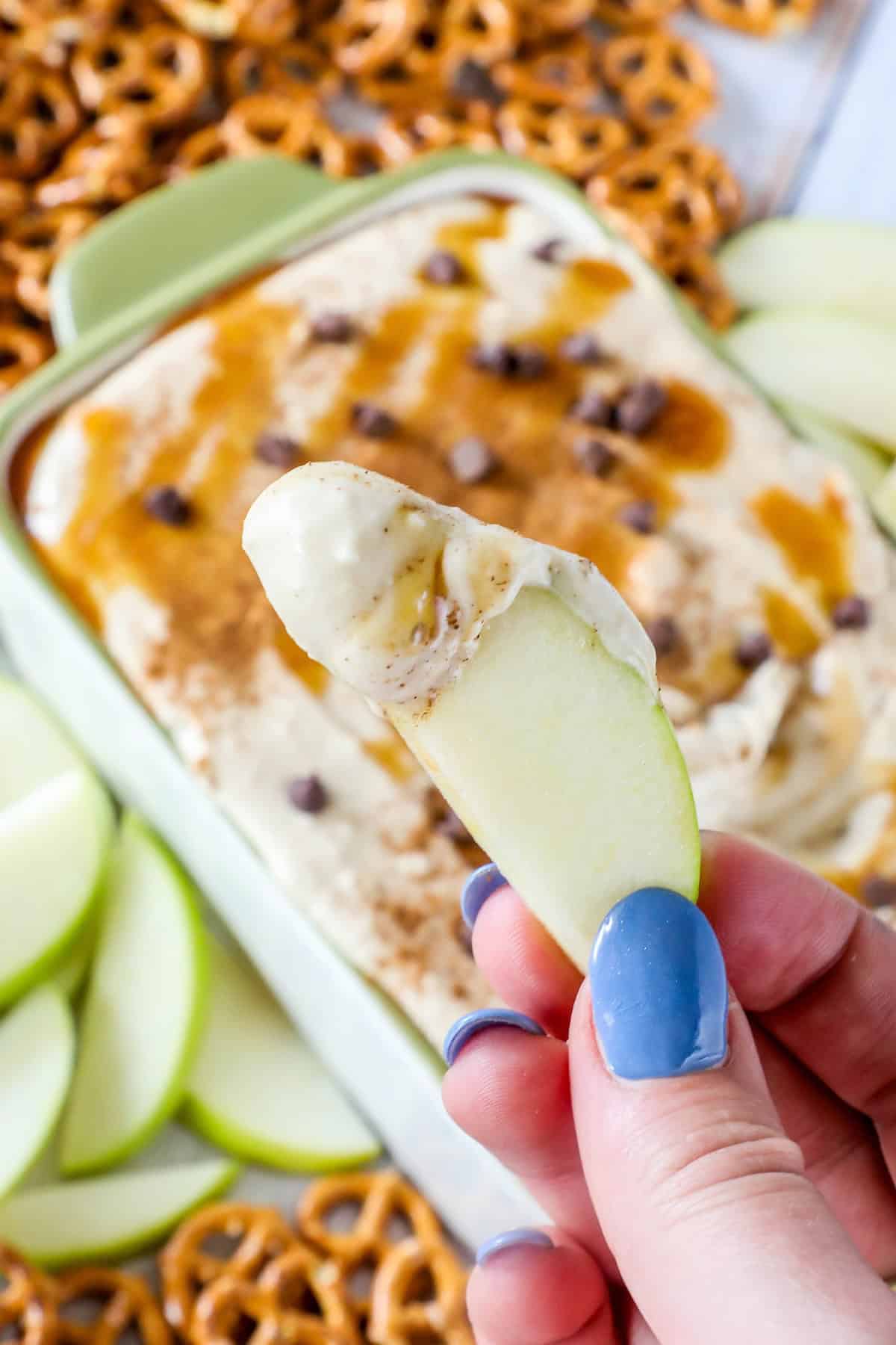 A person holding a slice of green apple dipped in a rich caramel apple dip, with pretzels and more apple slices visible in the background.