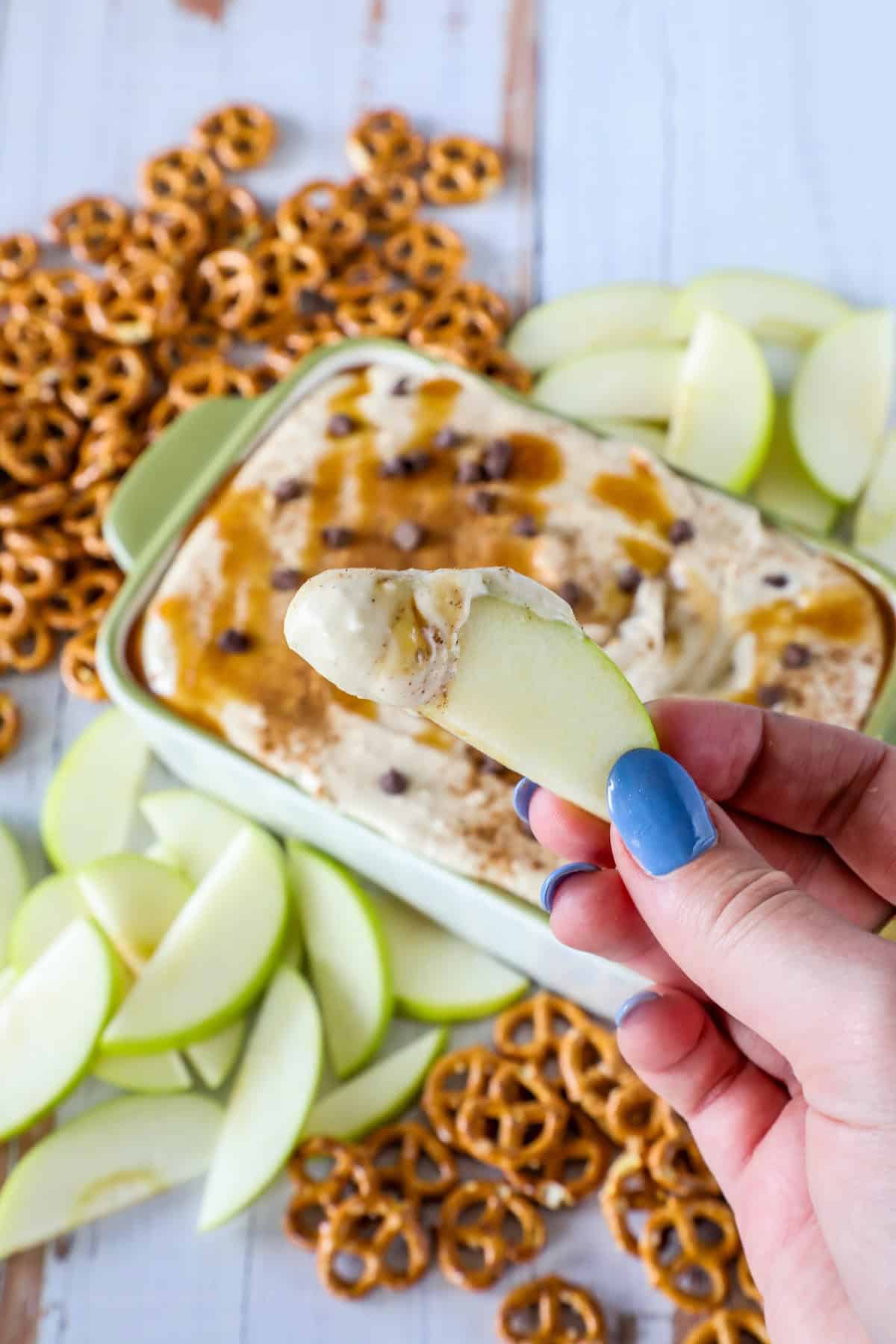 A hand holds a green apple slice dipped in rich caramel apple dip, surrounded by a tray of dip, pretzels, and more apple slices on a table.