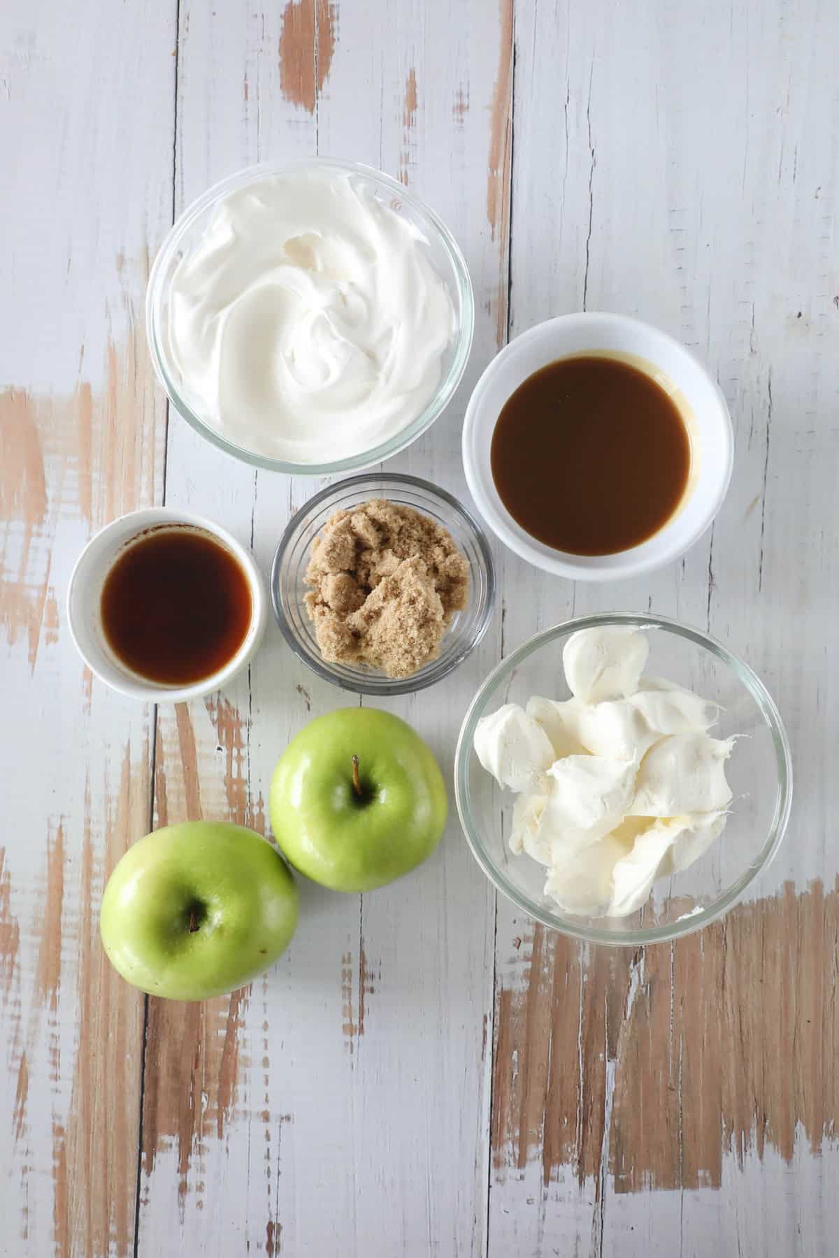 On a wooden surface, ingredients for a caramel apple dip include two green apples, bowls of brown sugar, cream cheese, rich caramel, vanilla extract, and fluffy whipped cream.