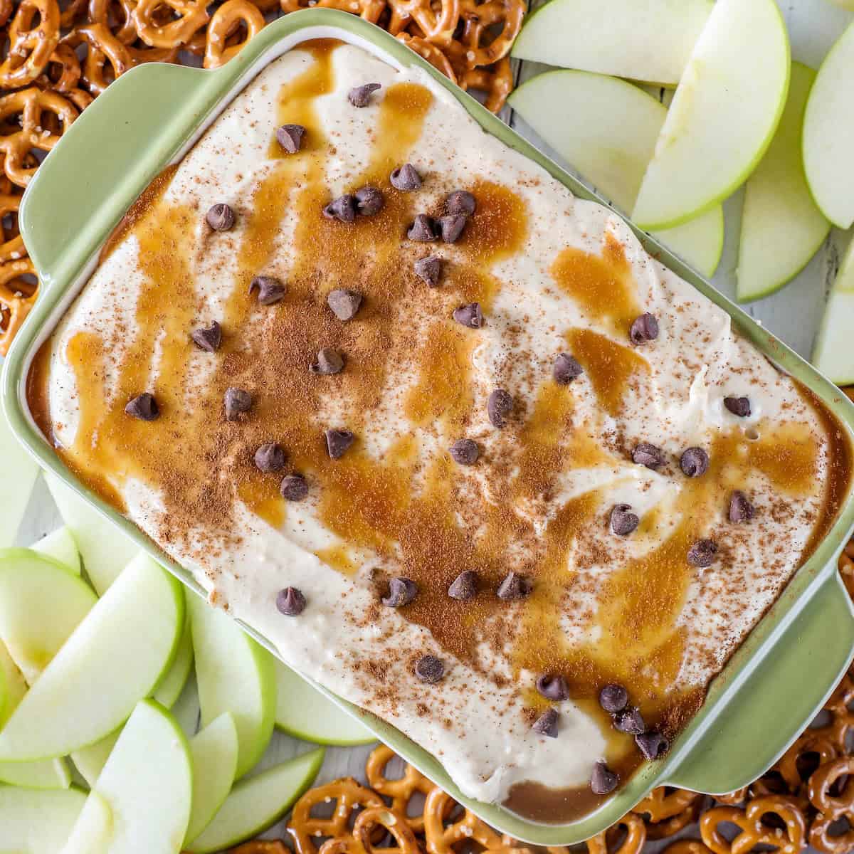 A delightful serving of caramel apple dip crowned with chocolate chips and cinnamon, encircled by crisp green apple slices and crunchy pretzels.