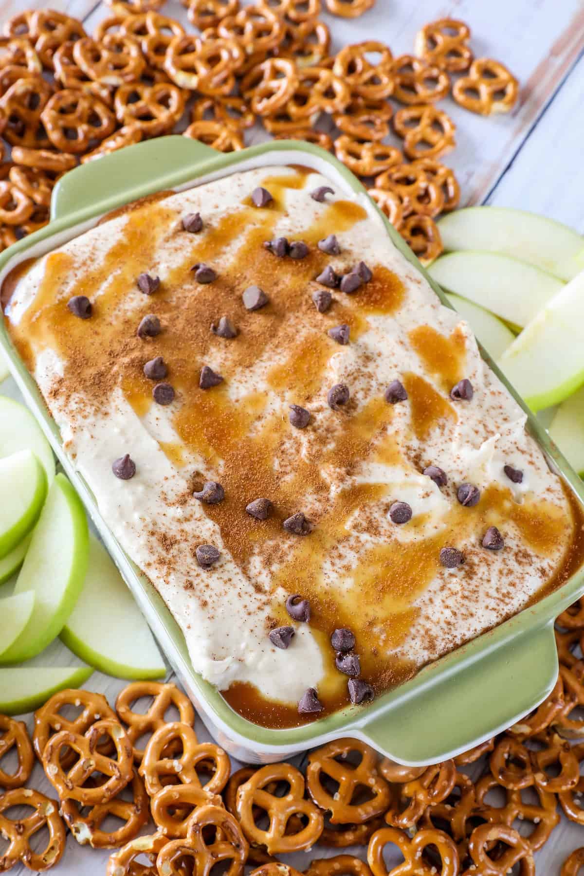 A green dish with creamy caramel apple dip topped with chocolate chips and cinnamon, surrounded by apple slices and pretzels.