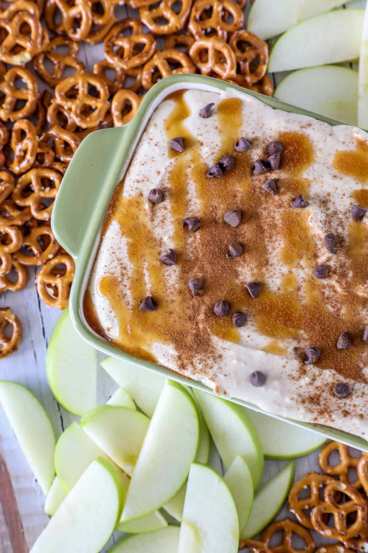 A tray of caramel apple dip, generously topped with chocolate chips and cinnamon, surrounded by crisp pretzels and sliced green apples.